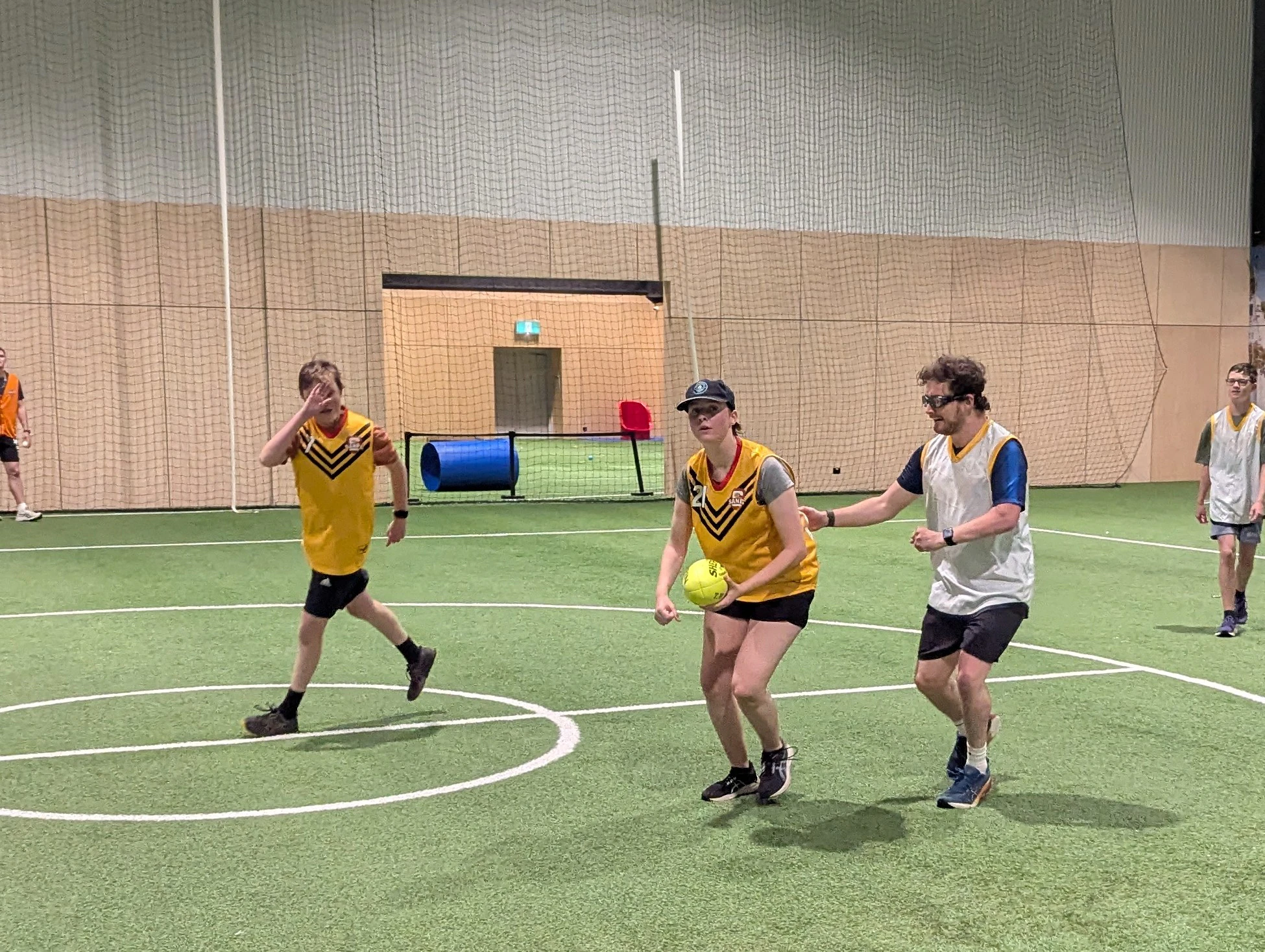 Indoor SANFL BLV Football game with players in yellow and white vests, one holding a bright yellow ball